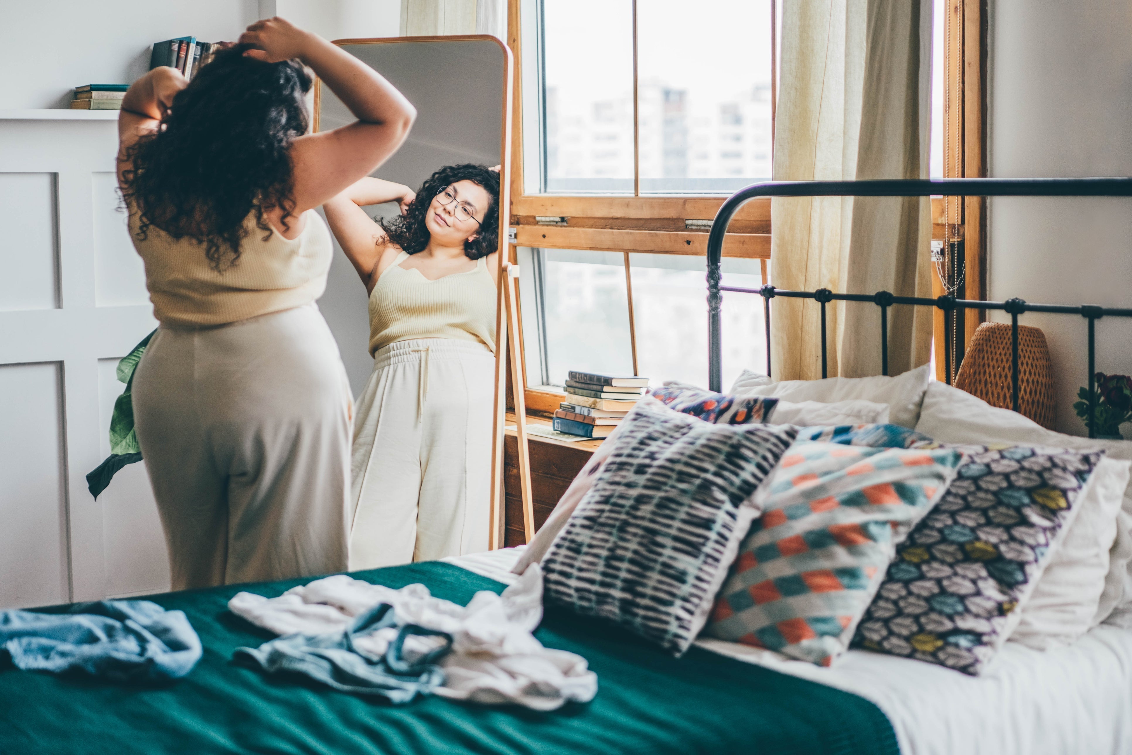 Woman standing in front of a mirror in a cozy bedroom, adjusting her hair while wearing a light yellow tank top and cream-colored wide-leg pants. A neatly made bed with colorful pillows and clothes laid out is in the foreground.