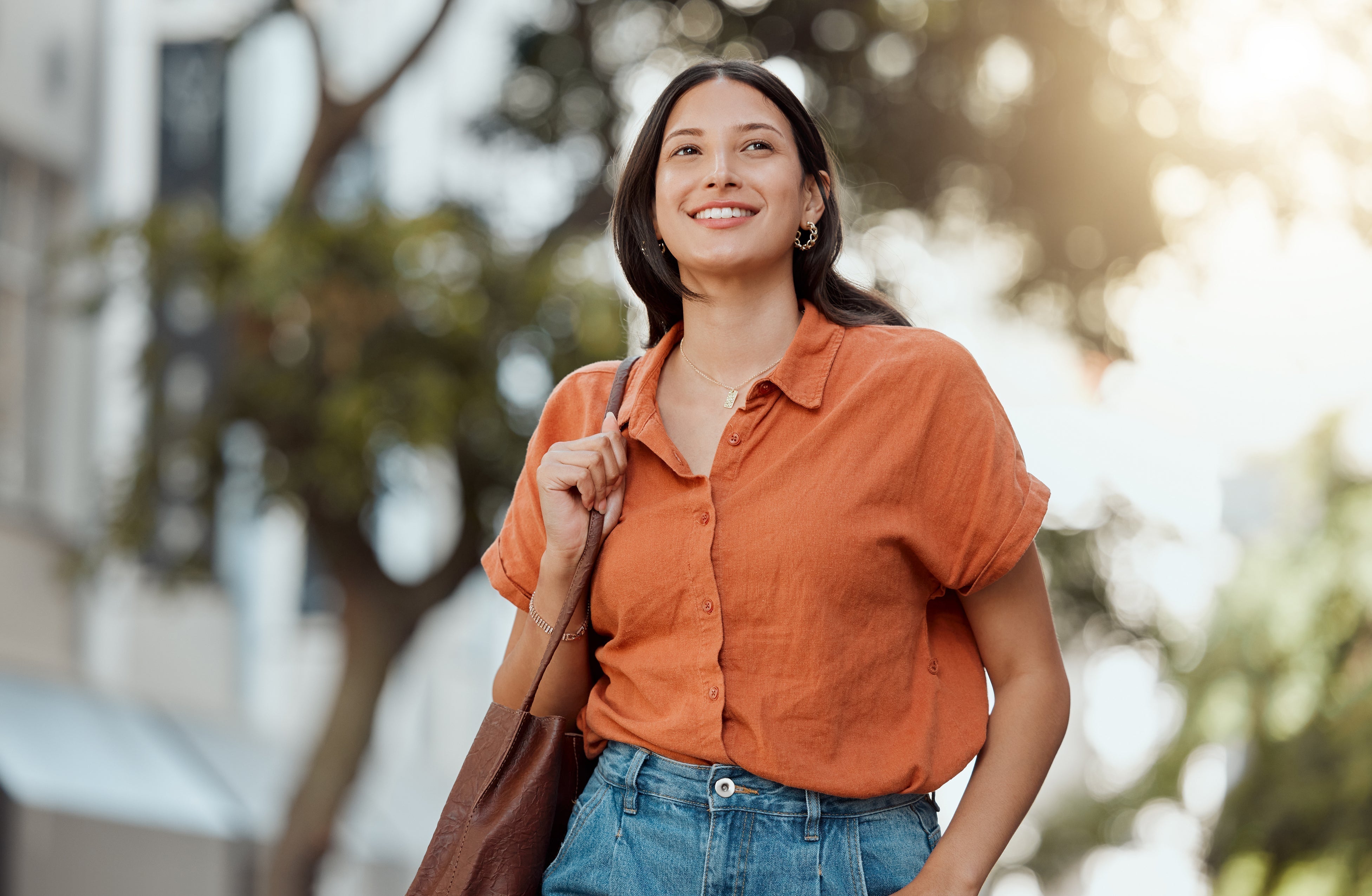 Confident young woman smiling while walking outdoors in a casual orange button-up shirt and high-waisted jeans, carrying a brown tote bag on her shoulder. Bright, sunny day with blurred trees and buildings in the background.
