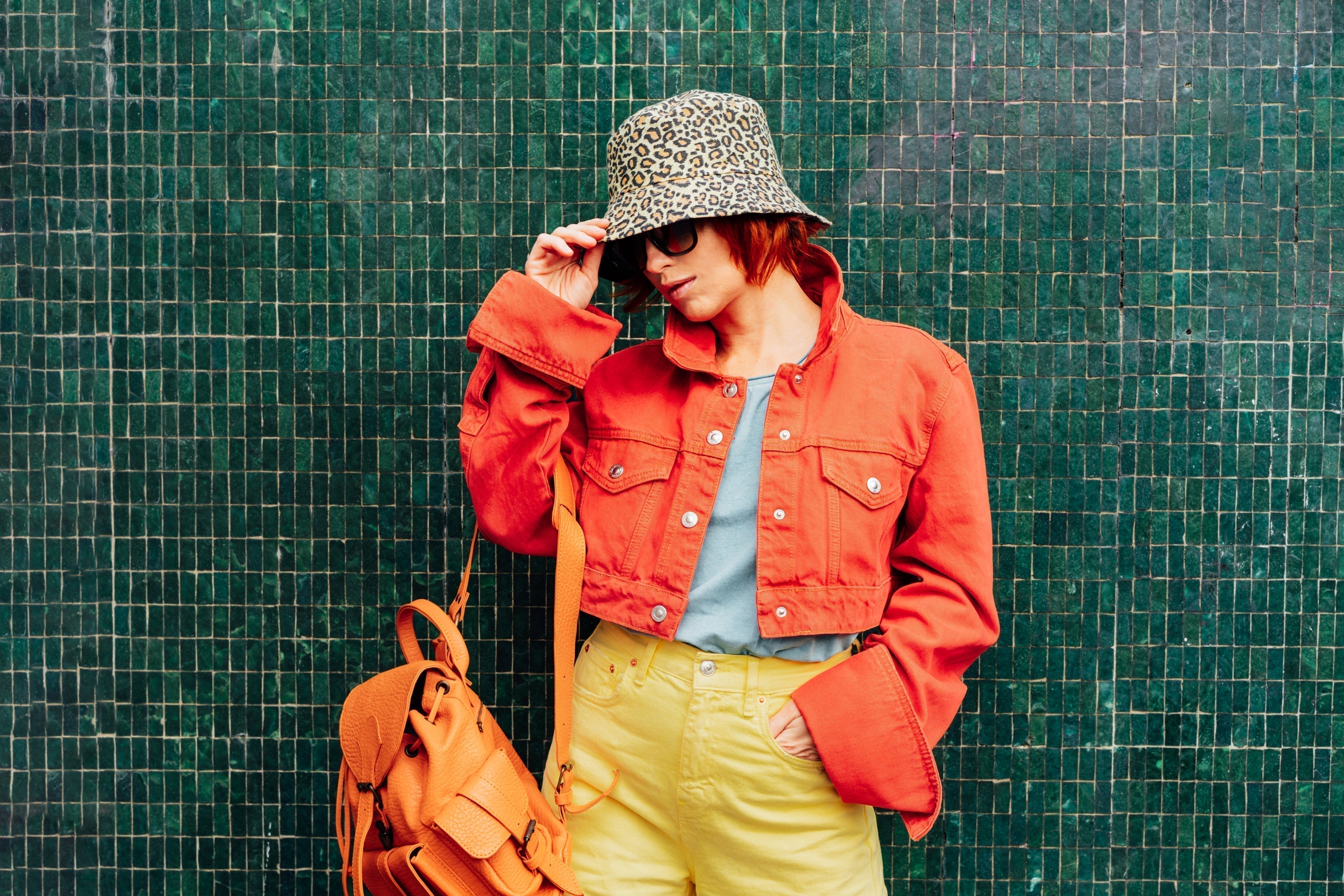 Stylish woman in a bold red cropped jacket, yellow high-waisted pants, and leopard-print bucket hat posing against a dark green tiled wall, accessorized with an orange leather backpack.