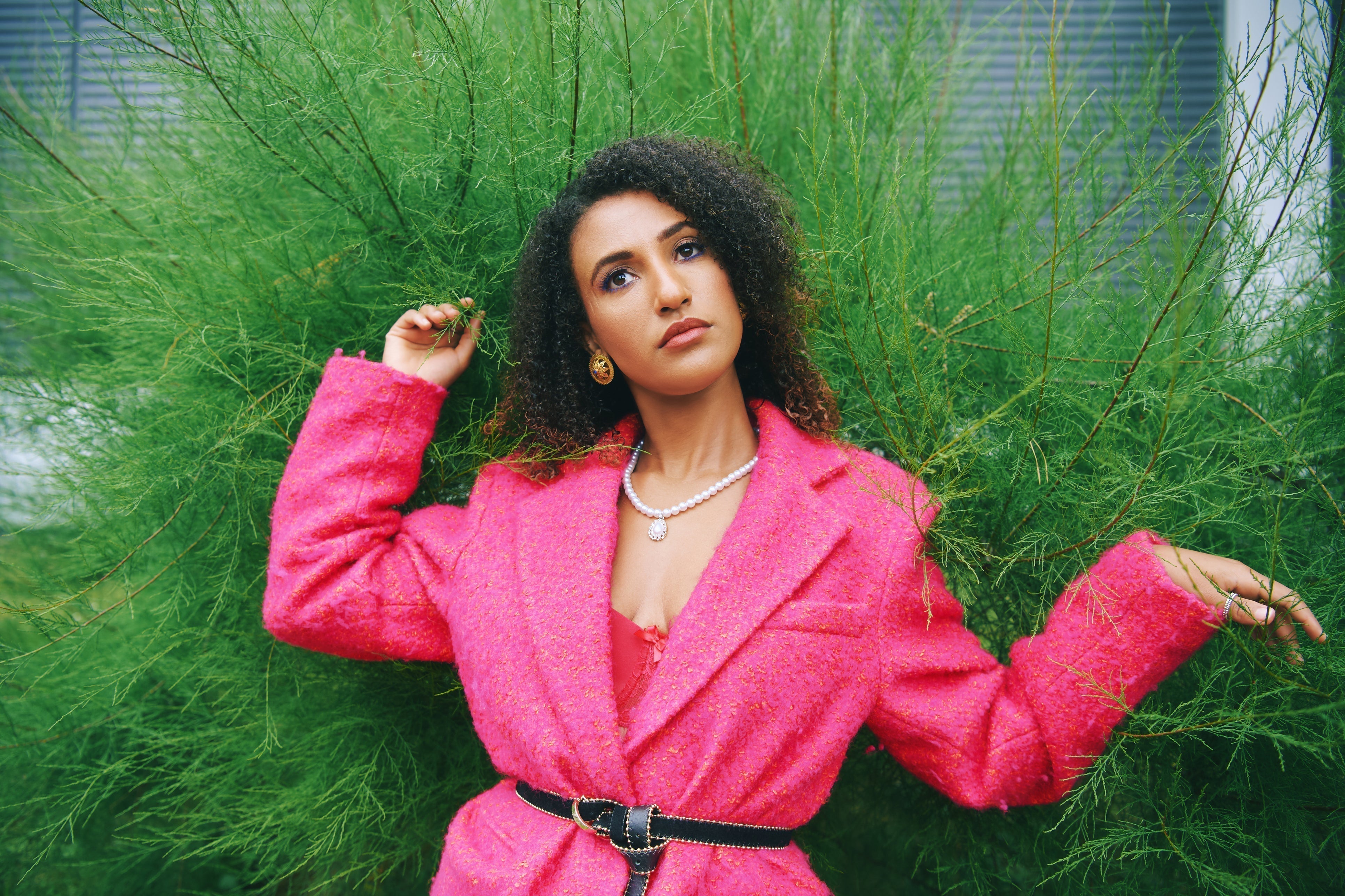 Woman wearing a bold hot pink textured coat with a black belt and pearl necklace, posing confidently in front of lush green foliage.