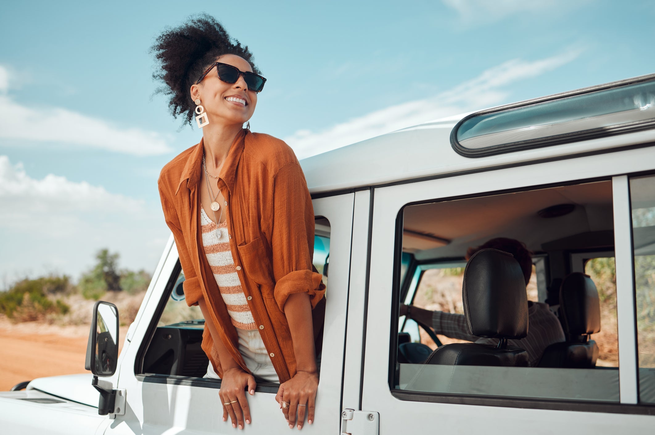 woman with striped ivory and orange shirt and a orange light blazer wearing sunglasses and earrings hanging out of a truck outside